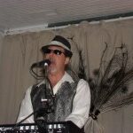 A musician in sunglasses and a fedora performs at a keyboard, with a decorative backdrop featuring peacock feathers.
