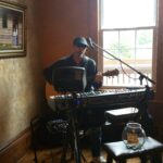 A musician plays guitar and keyboard in a cozy venue, with a jar for tips on a stool nearby.
