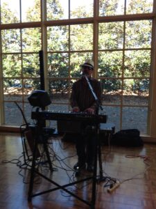 A musician performs at a venue with large windows, playing a keyboard and singing into a microphone.